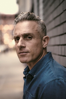 Miguel Luciano looks directly at the camera. He has very short and tidy hair. He is leaning against a grey brick wall, and wearing a denim button-down shirt. 
